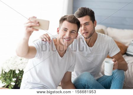 Let me take a selfie. Emotional young homosexual couple smiling and taking selfie while sitting on the sofa.