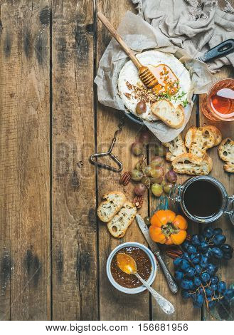 Homemade camembert cheese in small pan, honey, fig jam, persimmon, grapes, pecan nuts, grilled baguette slices and glass of rose wine over rustic wooden background, top view, copy space