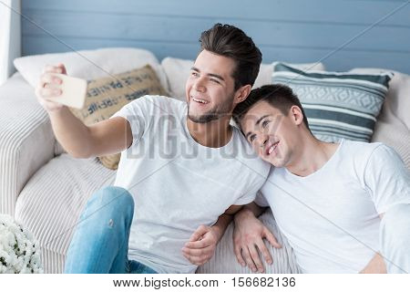 Fun time together. Happy handsome gay partners smiling and making selfies together while relaxing on a sofa.