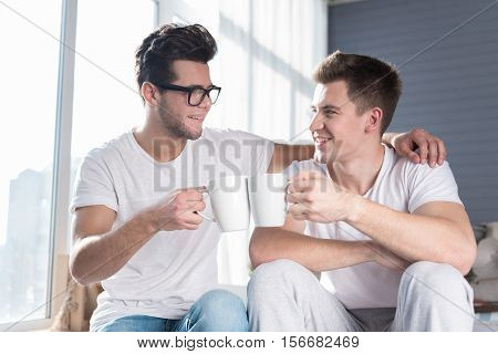 alternative relationships. Joyful young homosexual couple smiling and having a hot drink together while relaxing in an airy room.