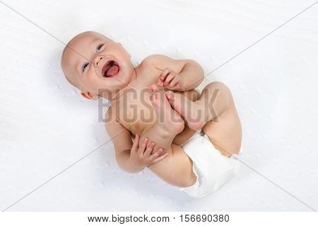 Funny little baby wearing a diaper playing on a white knitted blanket in a sunny nursery. Child after bath or shower on a fresh towel. Infant nappy change and skin care. Cute kid playing with his feet