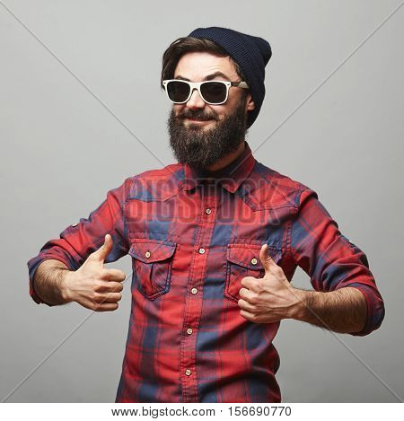 Smiling young man with long facial hair giving thumbs up sign. Happy hipster man with beard and sunglasses showing ok gesture.