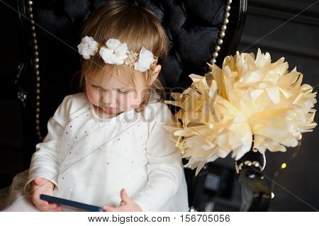 Bonny ruddy toddler sitting in the chair and enthusiastically looking at smartphone screen. In white dress with a wreath on his head the child like an angel.