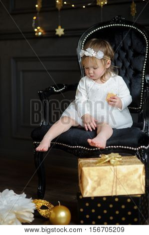 Little ruddy girl sits in a beautiful chair and eats cookies. Toddler is dressed in an elegant white dress. On the head of the girl a wreath. Room is festively decorated on floor boxes with gifts lie