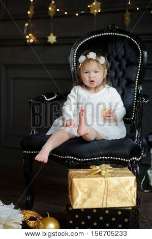 Little ruddy girl sits in a beautiful chair and eats cookies. Toddler is dressed in an elegant white dress. On the head of the girl a wreath. Room is festively decorated on floor boxes with gifts lie