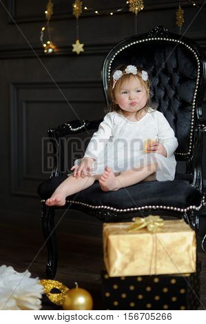 Little ruddy girl sits in a beautiful chair and eats cookies. Toddler is dressed in an elegant white dress. On the head of the girl a wreath. Room is festively decorated on floor boxes with gifts lie