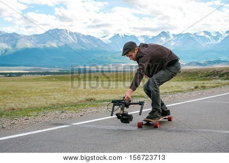 Man rides a longboard and shoots camera with electronic steadycam. Long road in the field on a background of mountains and glaciers. Altay, Russia.