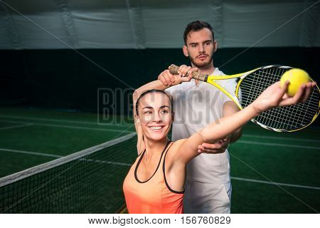 Engaged in process. Cheeful beautiful woman serving a ball while professional instructor controlling the process while training in an indoor court