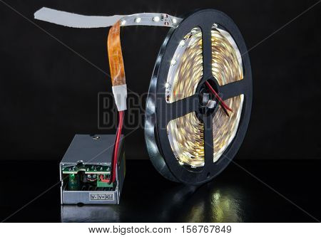 Close-up of luminous LED Light strip wound around a bobbin and a voltage converter on a black background.
