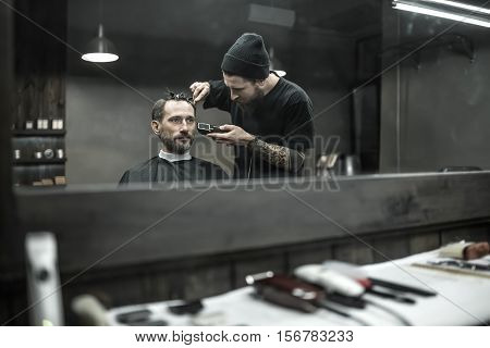 Peerless barber with a beard and a tattoo is cutting the hair of his bearded client in the barbershop. He is using a cutting comb and a hair clipper. Customer has hairgrips on the head. Horizontal.