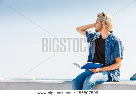 Holiday outdoor leisure time introvert relaxation concept. Sitting hipster man wearing jeans outfit having break from reading book outside on sunny day sea in background