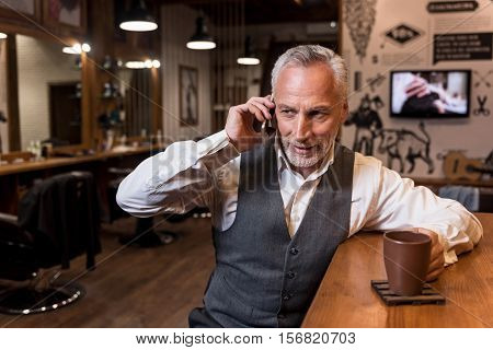 Business during coffee break. Portrait of attractive senior businessman sitting at bar counter and having mobile conversation.