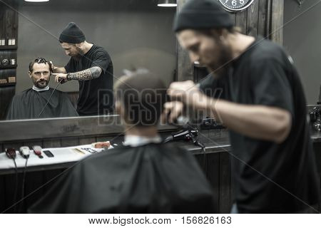 Matchless barber with a beard and a tattoo is cutting the hair of his bearded client in the barbershop. He is using a cutting comb and a hair clipper. They both reflected in a mirror. Horizontal.