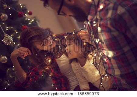 Beautiful young family enjoying their holiday time together decorating Christmas tree arranging the christmas lights and having fun