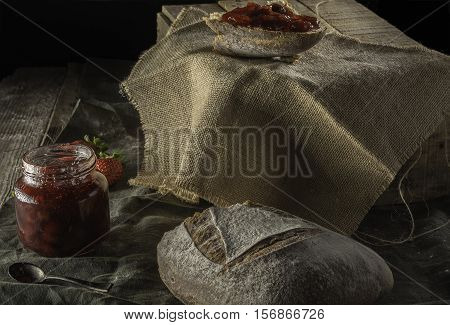 Piece of bread strawberry jan on top of a wooden box with a jar of strawberry jam with a piece of country bread and a small spoon.