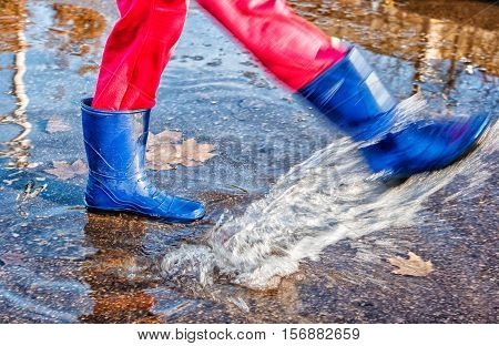 girl in blue boots and red pants standing in a puddle of water splashes with fallen yellow leaves on autuman day closeup