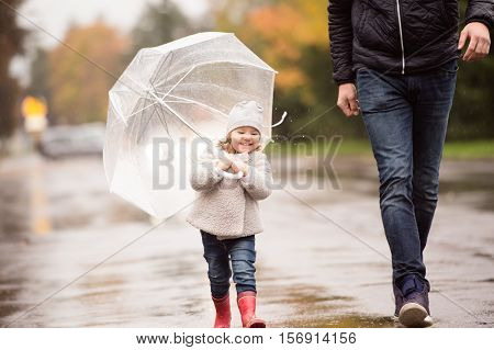 Unrecognizable young father with cute little daughter under the umbrella. Walking in the street on a rainy day.