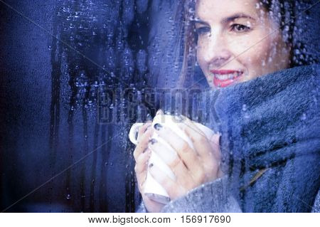 attractive adult woman drinking coffee and looking out of the window on rainy day, close up portre in winter time