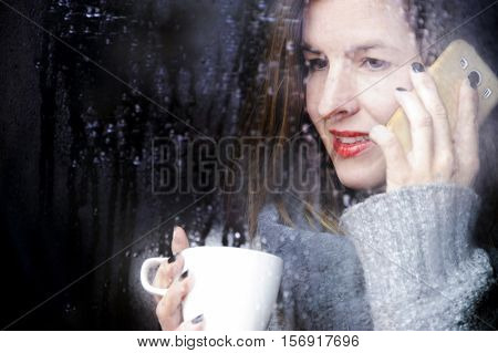 attractive adult woman drinking coffee speaking with mobile and looking out of the window on rainy day, close up portre in winter time