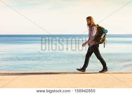 Man hiker backpacker walking with backpack by seaside at sunny day. Adventure summer tourism active lifestyle. Young long haired guy tramping
