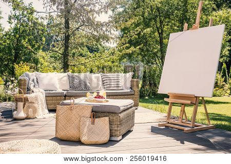 Mockup Of Easel On Wooden Terrace With Rattan Table And Settee In The Garden. Real Photo