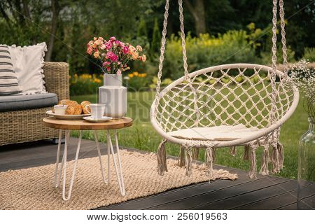 Hanging Chair Next To Table With Flowers On Terrace In The Garden During Spring. Real Photo