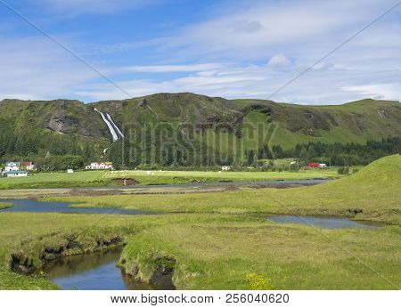 Rural Landscape With Colorful Farm Houses, Blue River Stream On Green Grass Meadow With Spruce Tree 