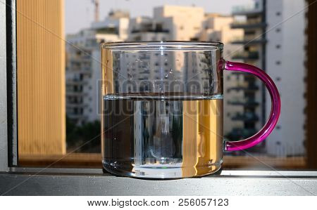 A Glass Mug With A Colored Handle, Standing In The Window Aperture.