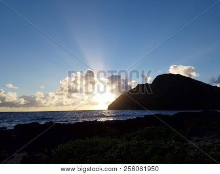 Ocean Waves Lap On Makapuu Beach As Sunrises Through The Clouds With Makapuu Lighthouse And Point In