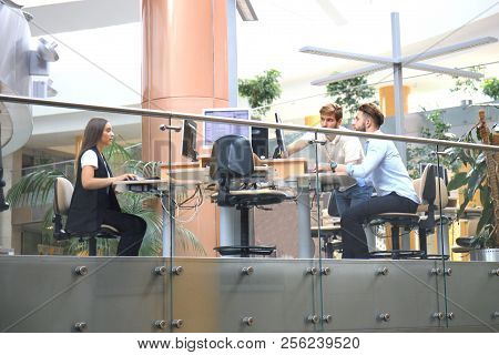 Group Of Young People In Casual Wear Sitting At The Office Desk And Discussing Something While Looki