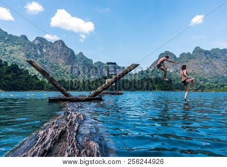 Two Teenage Kids Jumping In The Lake With Mountains And Rain Forest In The Background During A Sunny