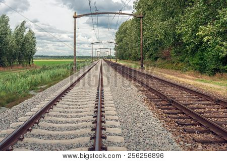 Dutch Landscape With Rusty Train Tracks With Overhead Lines. It Is A Cloudy Day In The Dutch Summer 