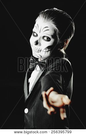 Portrait of a child boy with carnival make-up of a skeleton wearing elegant tail-coat. Studio shot over black background. Halloween. Dia de los muertos. The Day of the Dead.