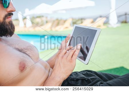 Cut View Of Bearded Guy Sits On Sunbed And Wears Glasses. He Holds White Tablet And Looks At It. He 