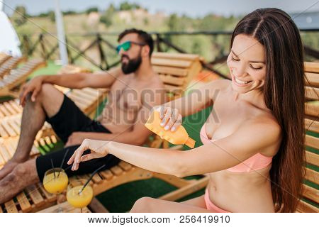 Nice And Positive Girl Sits And Puts Some Sunproof Cream On Skin. She Holds Orange Bottle In Right H