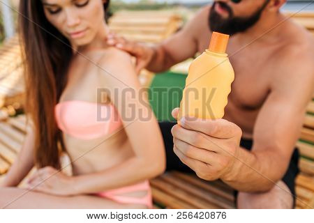 Serious Guy And Girl Sit. He Puts Some Sunproof Lotion From Orange Bottle On Her Shoulders And Back.