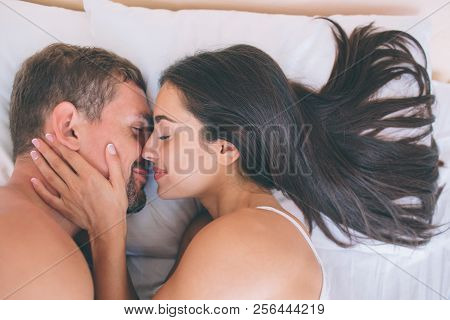 Cheerful And Nice Couple Is Lying In Bed Close To Each Other. Girl Holds Her Hand On His Neck. They 