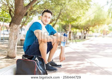 Tired Male Runner Holding Waterbottle While Sitting On Footpath In Park