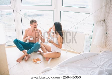 Girl And Guy Are Sitting On Floor With Their Legs Crossed. They Eat Croissans And Drink From White C