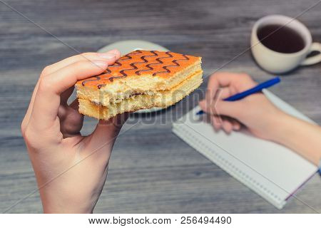 A Close Up Of Hand Holding Bitten Sponge Cake. Background Of Writing Hand And Cup O F Hot Coffee On 