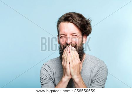 Joyful Excited Happy Smiling Man Covering Mouth With Hands. Portrait Of A Young Bearded Guy On Blue 