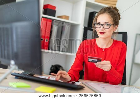 A Young Girl Is Sitting At The Desk In The Office, Holding A Bank Card In Her Hand And Typing On The