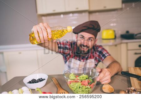 Nice And Positice Chief Stands In Kitchen At Table And Holds Bottle Of Oil. He Pours Some Of It Into