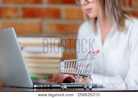Young Businesswoman Sitting On A Table With Laptop Computer And Books And Supermarket Cart On Forego