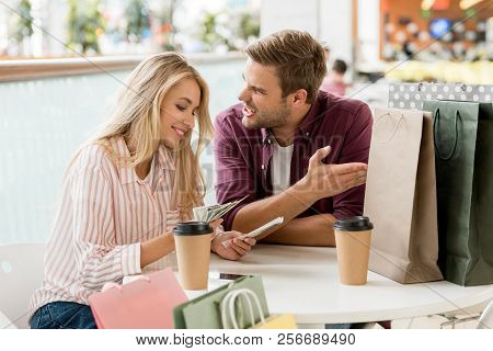 Smiling Young Woman Counting Cash Money After Shopping While Her Upset Boyfriend Sitting Near At Tab