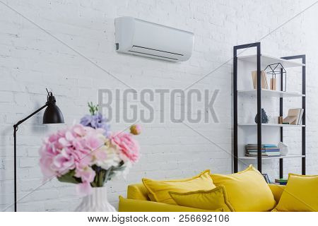 Interior Of Modern Living Room With Flowers In Vase, Yellow Couch And Air Conditioner Hanging On Whi