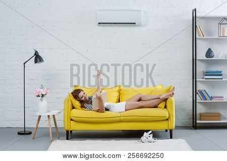 Relaxed Young Woman Reading Book On Sofa And Pointing At Air Conditioner With Remote Control