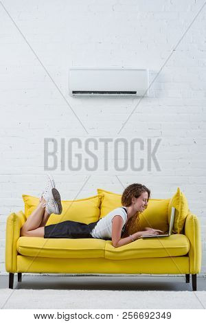 Happy Young Woman Working With Laptop While Lying On Couch Under Air Conditioner Hanging On Wall
