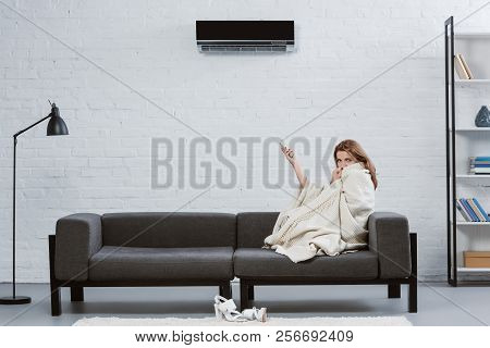 Young Woman Covered With Blanket On Couch Under Air Conditioner Hanging On Wall