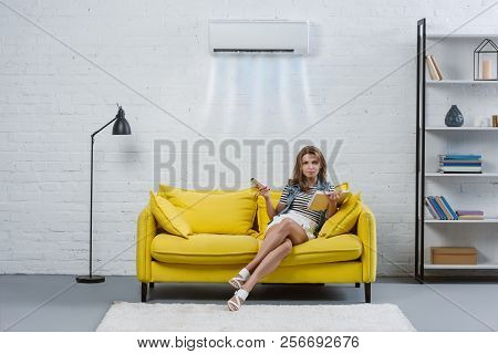 Beautiful Young Woman With Book Sitting On Couch And Pointing At Air Conditioner Hanging On Wall Wit